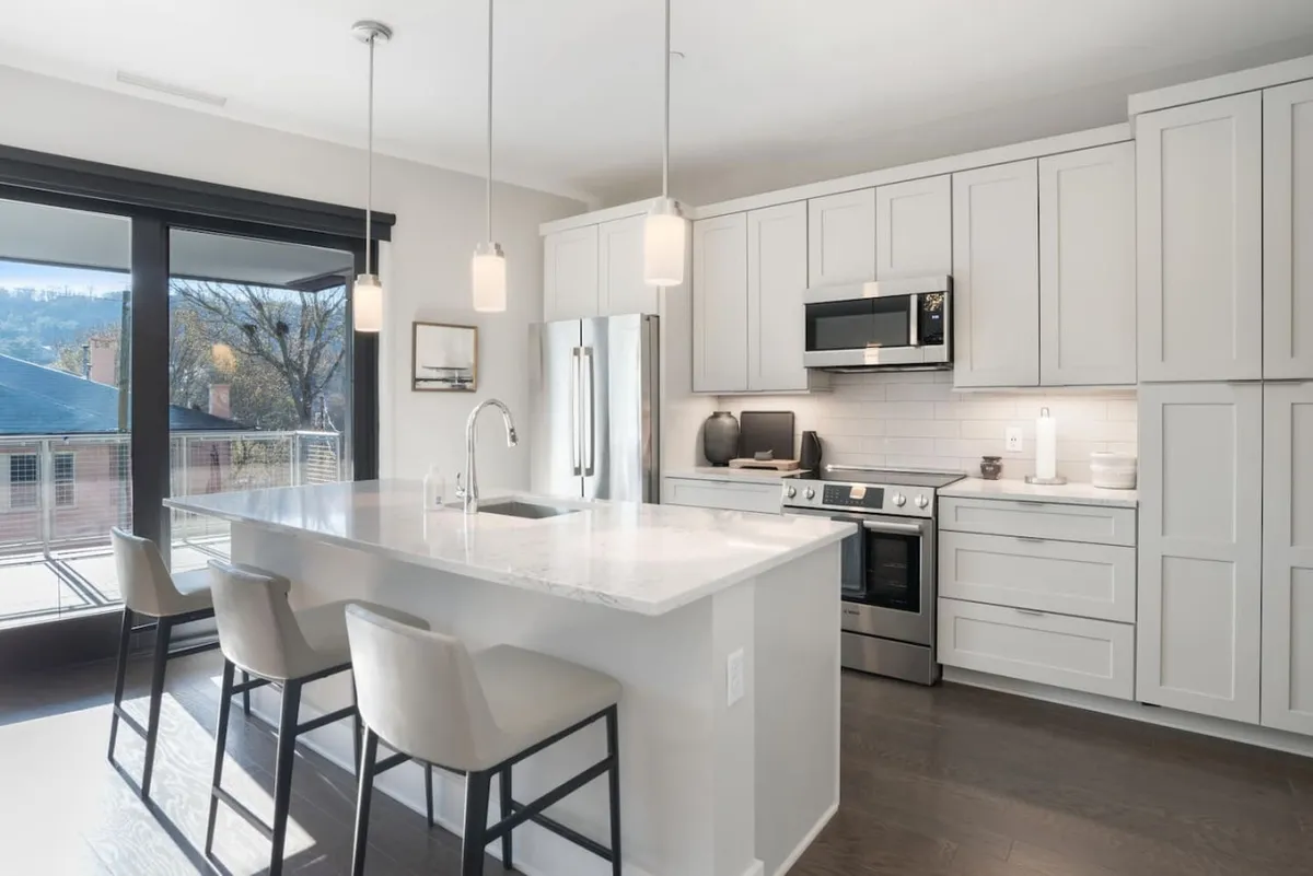 A bright, contemporary kitchen featuring a large island with minimalist cabinetry, stainless steel appliances, and expansive windows that flood the space with natural light. The scene captures both the functional and social aspects of the kitchen, with plenty of room for cooking and casual gatherings.