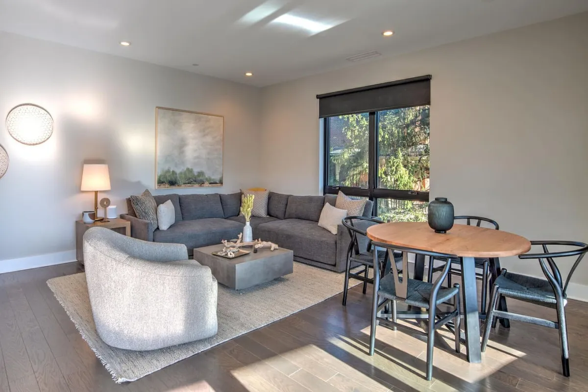 A stylish, open-concept living area featuring a modern sofa, eye-catching contemporary artwork on the wall, a dining table in the background, and large east-facing windows with city views. The space feels bright and airy, ideal for relaxing or socializing.