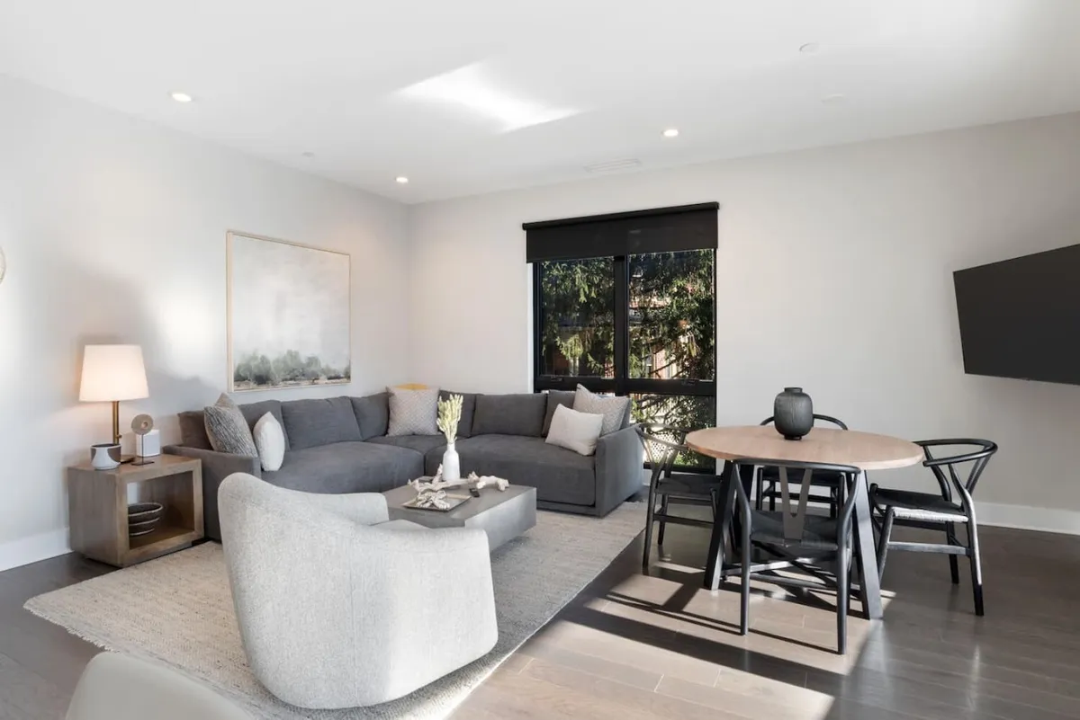 A cozy living space with a large gray sectional sofa, round dining table, and prominent wall art. A large window frames greenery outside, creating a peaceful yet vibrant atmosphere suitable for gatherings or quiet evenings.