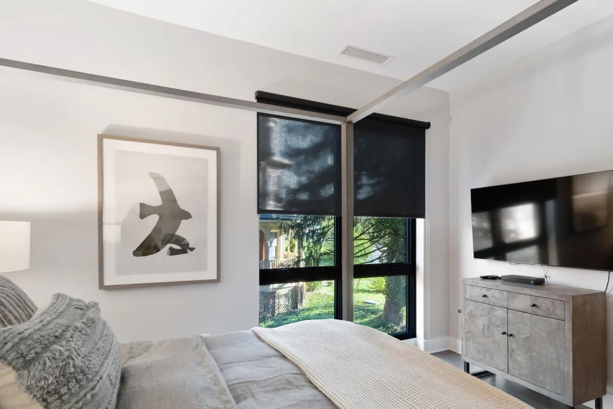 A peaceful bedroom with a comfortable queen bed dressed in textured bedding, a TV unit with a television for relaxing evenings, expansive windows with blinds for adjustable privacy, and modern wall art. The room feels inviting and organized, suggesting generous closet space even if not visible.