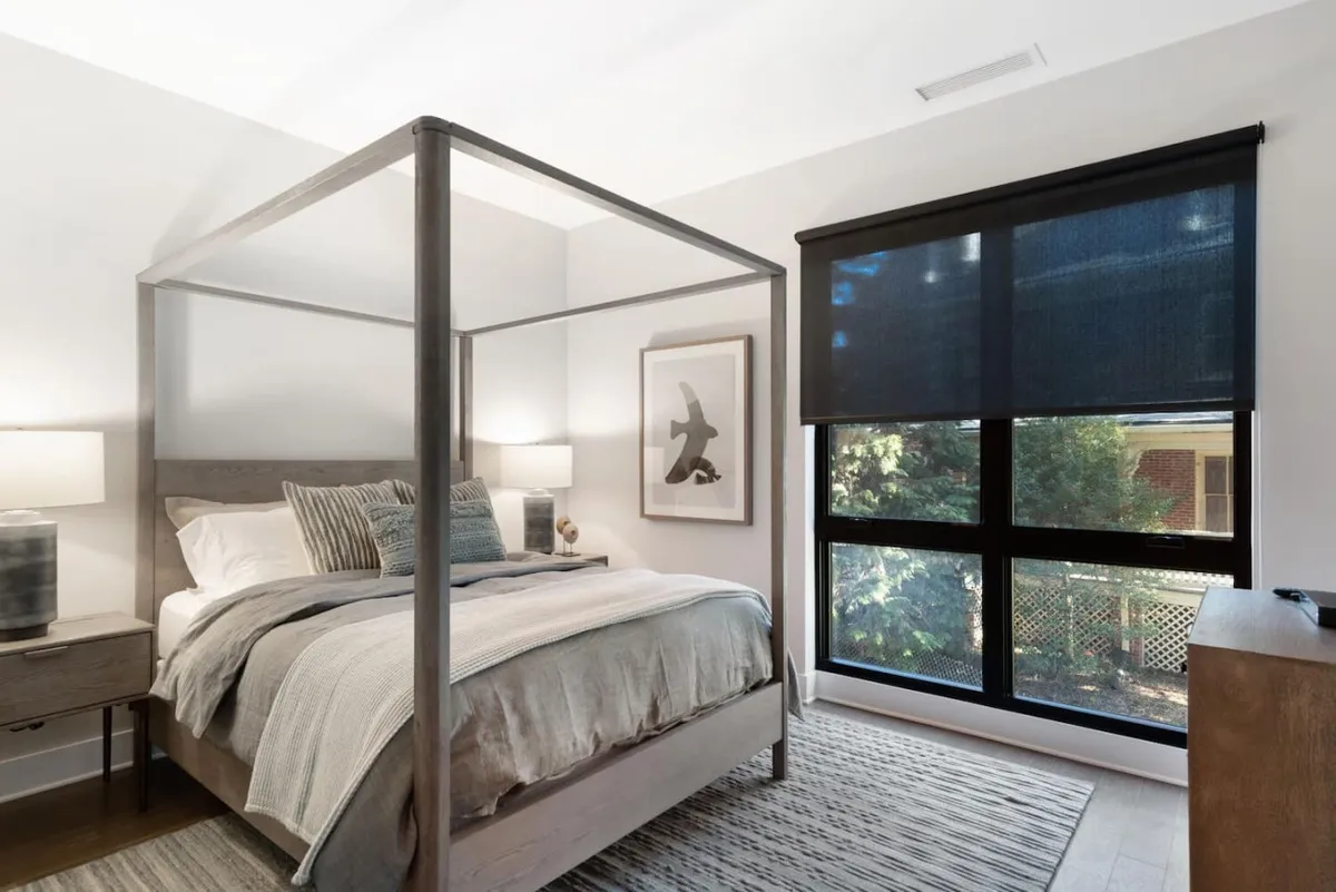 A serene bedroom featuring a plush canopy queen bed, soft linens, a nightstand with a lamp, a window fitted with black shades for privacy, and tasteful framed artwork. The room exudes a cozy and tranquil ambiance, inviting relaxation and rest.