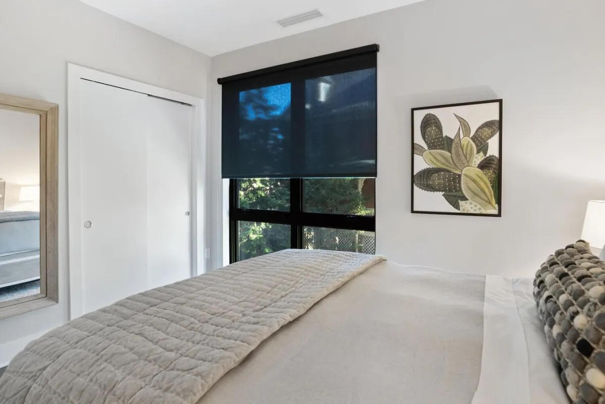 A tranquil bedroom with a textured quilt bed, a large window with adjustable blinds, calming wall art, and a full-length mirror. The scene is bathed in natural light, showcasing a comfortable and restful space ideal for business travelers seeking privacy and relaxation.