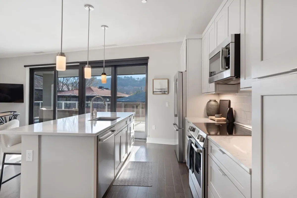 A bright, open-concept kitchen and living area featuring a spacious island countertop, modern appliances, and expansive windows that fill the space with natural light. The windows hint at the exterior cityscape, subtly connecting the interior with the urban environment outside.